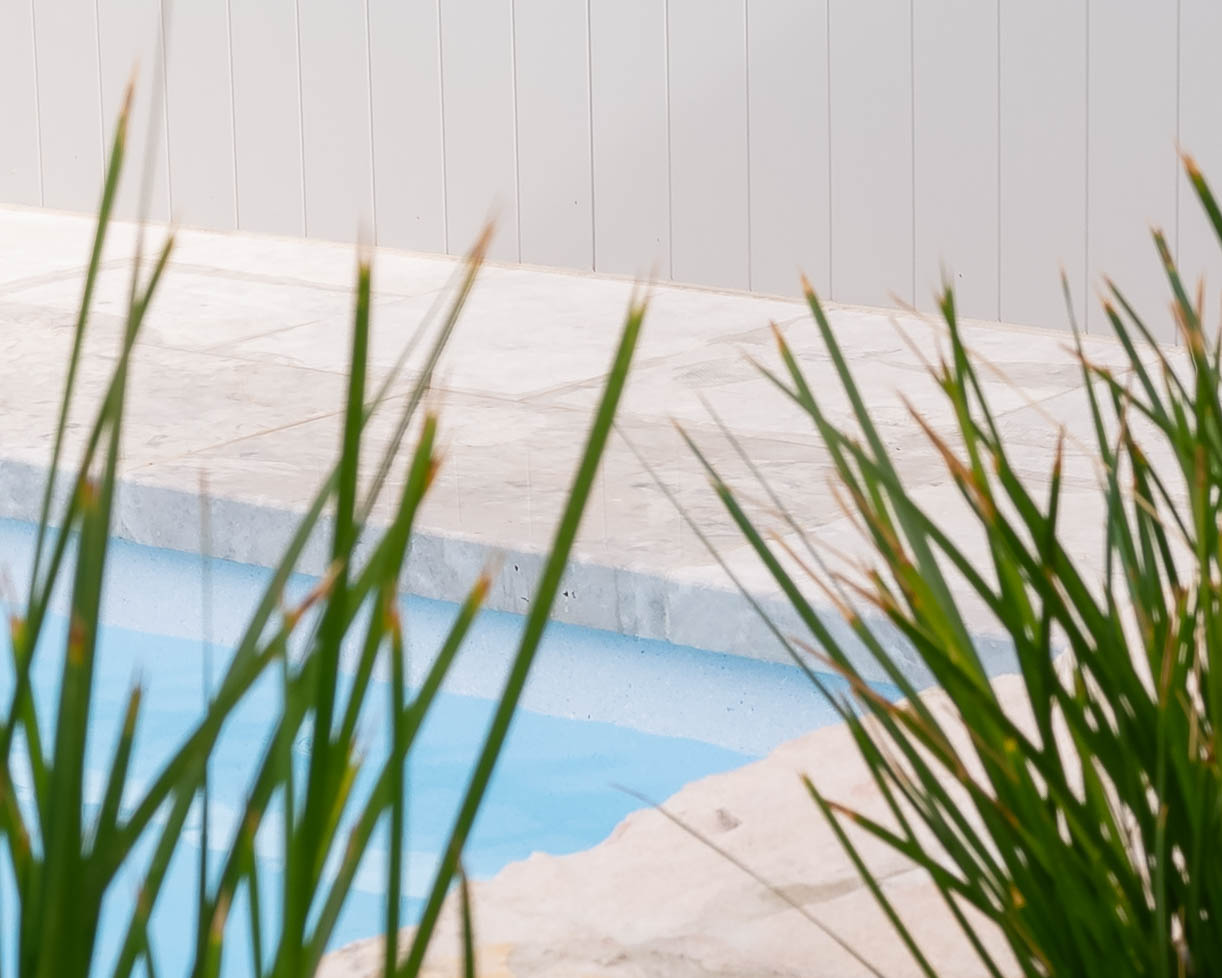 Close-up of honed travertine pool tiles meeting tropical foliage, showcasing seamless outdoor design in a Melbourne residence.